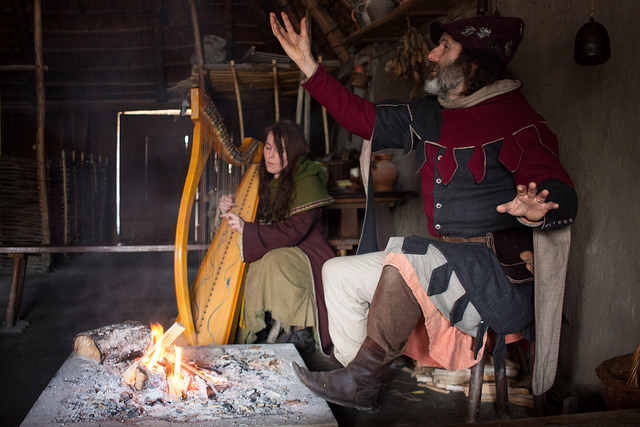 Man in medieval outfit telling a story with harpist in the background