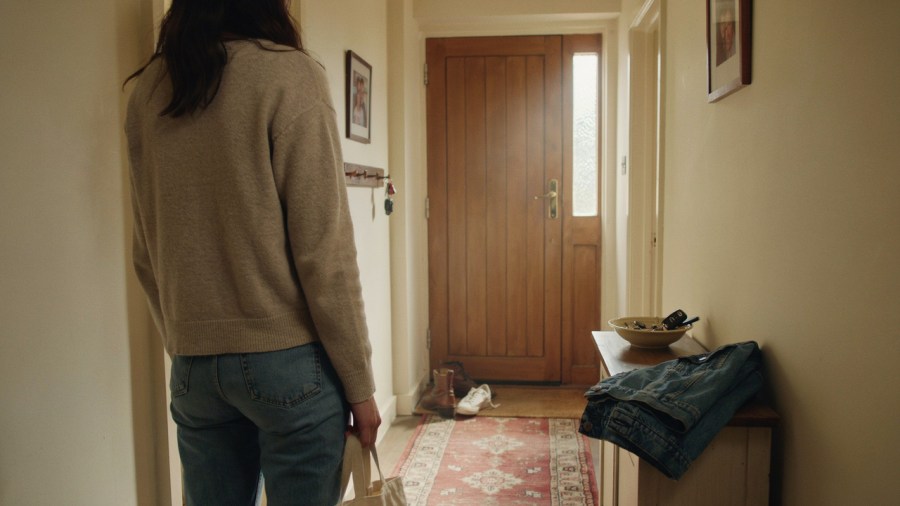 Over the shoulder photograph of a young woman in the narrow front hall of a house, the wooden front door at one end of it is closed and she has her hand on the door handle, ready to open it and leave Ideogram image