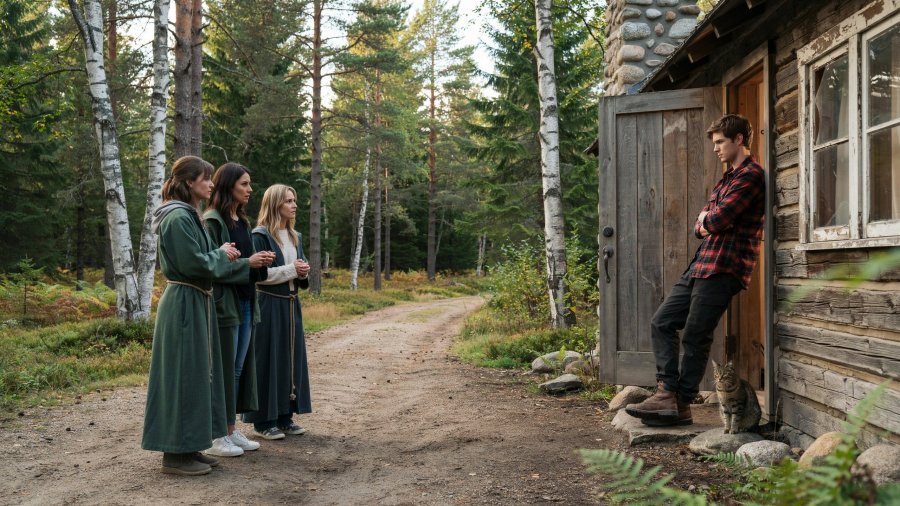 Photo of a small group of worried-looking women, with green priestess robes over casual modern clothing, standing outside a wood cabin on a small hill in a forest clearing, with a dirt road leading in, talking to a bored-looking man in the doorway of the cabin Ideogram image