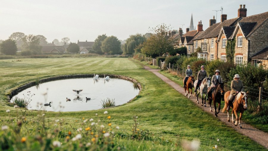 Cinematic photo of a large English village green with a pond in the middle, with ducks and two swans paddling in it. A group of five horses, ridden by older women walk along a narrow dirt track on the edge of the green, parallel to the road, with houses on the other side Ideogram image